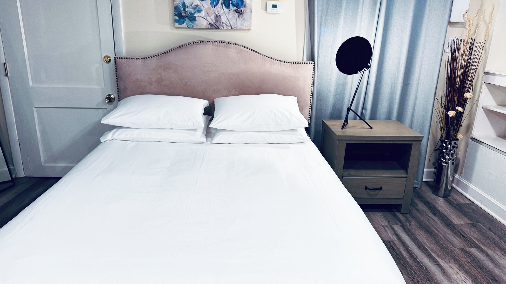 Ein ordentlich bezogenes Bett mit weißen Leinentüchern, ein Kopfteil mit dekorativer Verzierung, ein Nachttisch mit einer Lampe und ein an der Wand hängendes Kunstwerk.