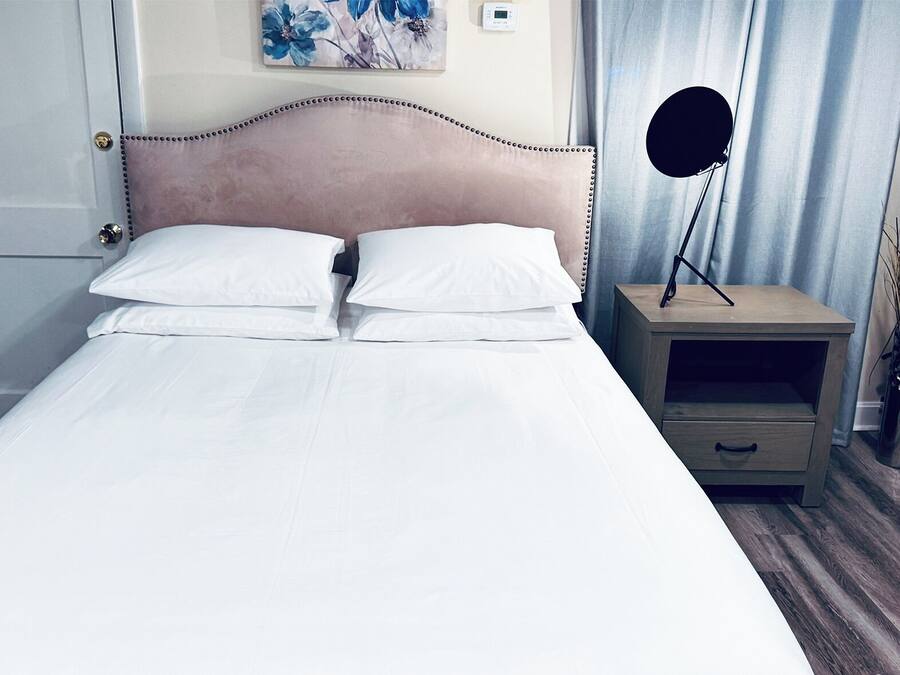 Ein ordentlich bezogenes Bett mit weißen Leinentüchern, ein Kopfteil mit dekorativer Verzierung, ein Nachttisch mit einer Lampe und ein an der Wand hängendes Kunstwerk.