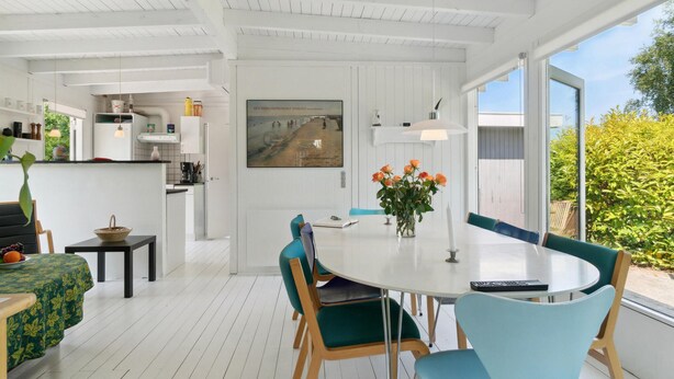 A modern kitchen with white cabinetry, a dining area with a round table and teal chairs, and a large window offering a view of greenery.
