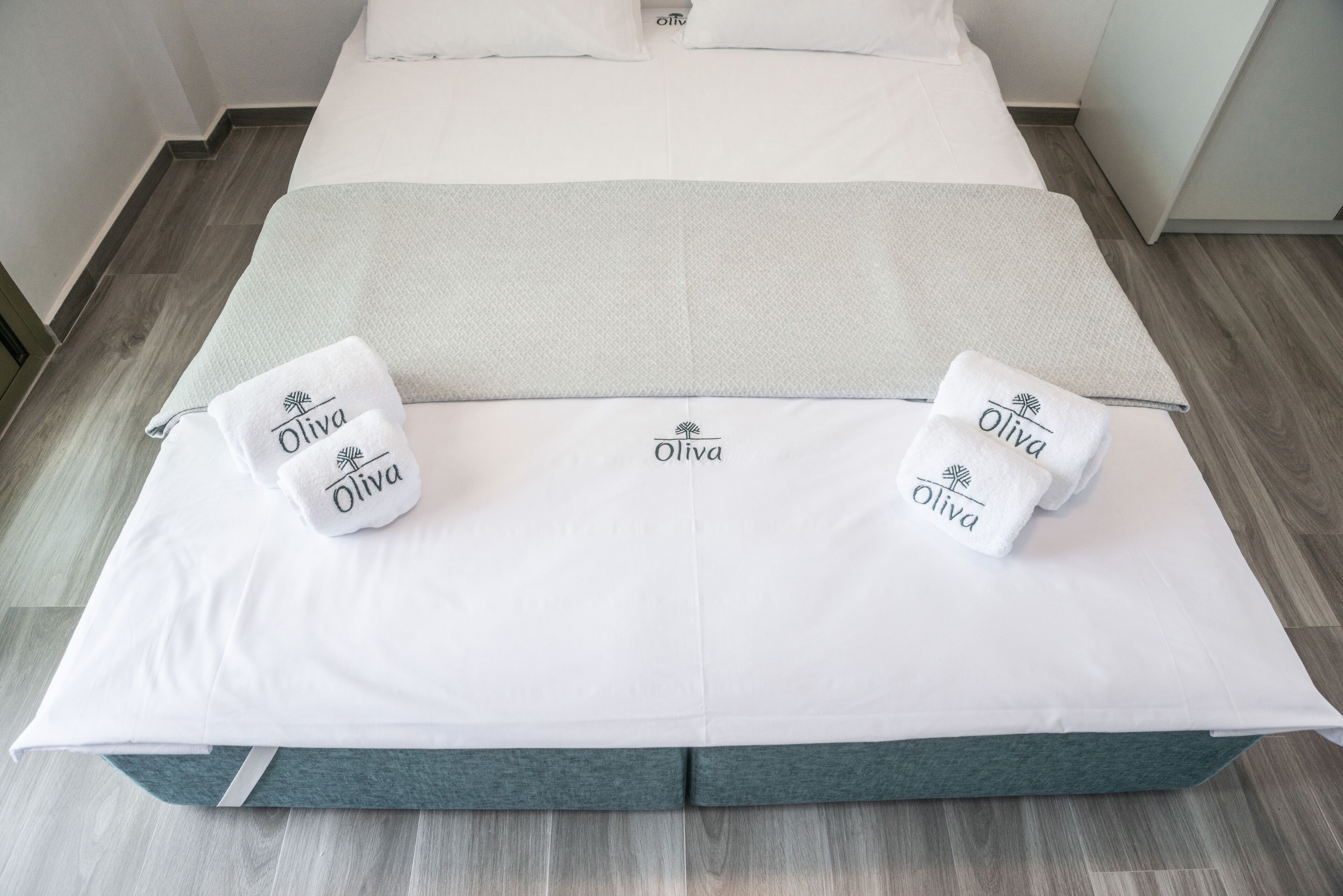 A neatly made bed with a white and teal bedspread, two towels with the name "Oliva" on them, and a logo on the bedspread.