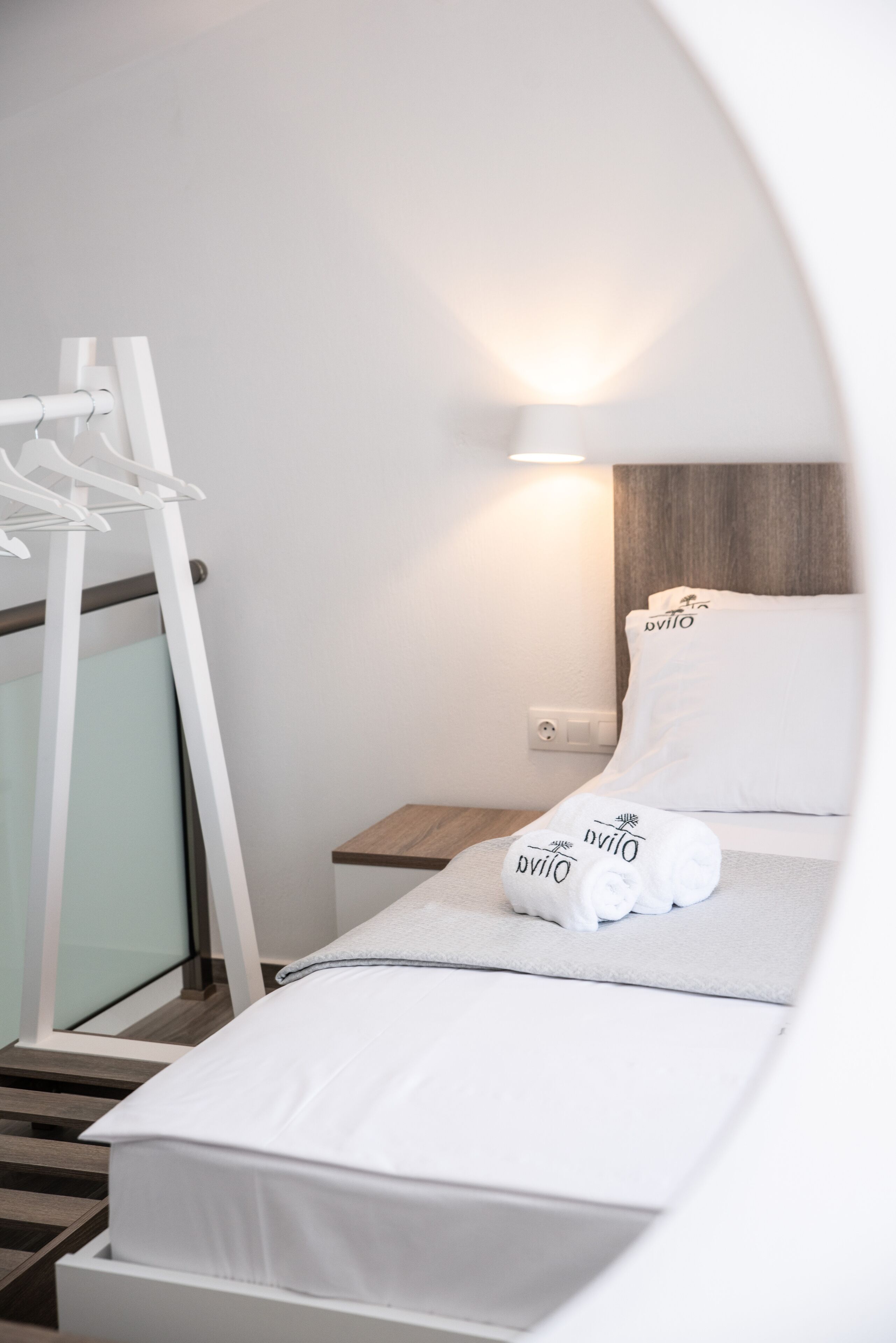 A neatly made bed with white linens and pillows, a wooden nightstand, a white towel, and a wall-mounted light fixture.