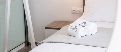 Una cama bien hecha con sábanas y almohadas blancas, una mesita de noche de madera, una toalla blanca y un aplique en la pared.
