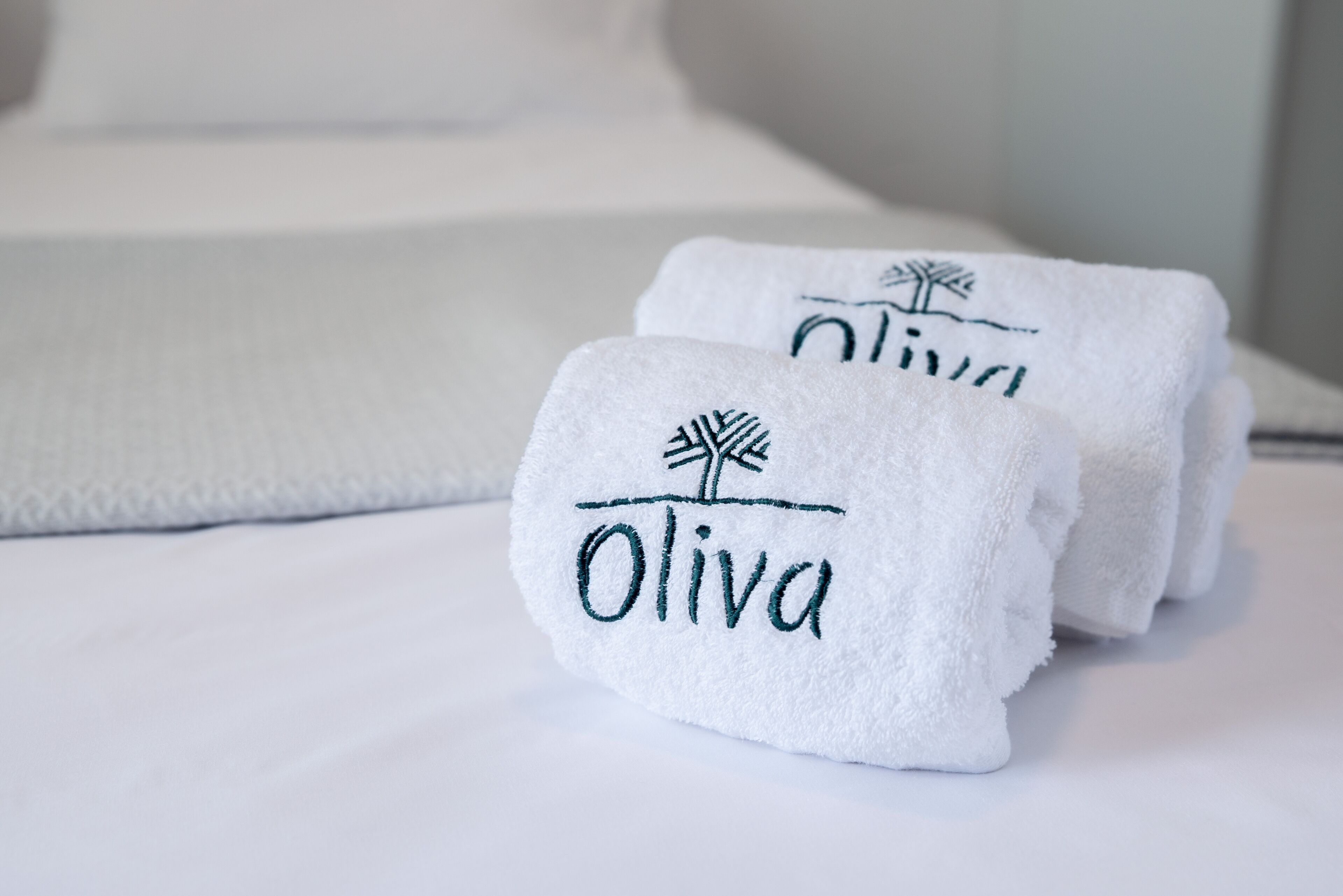 Zwei ordentlich gefaltete Handtücher mit dem Schriftzug "Oliva" und einem Palmenmotiv auf einem Bett.