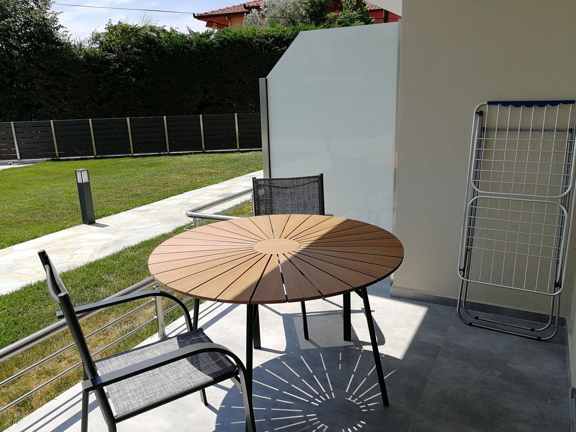 Un patio exterior con una mesa redonda y dos sillas, una parrilla de metal y un camino que lleva a una zona verde.