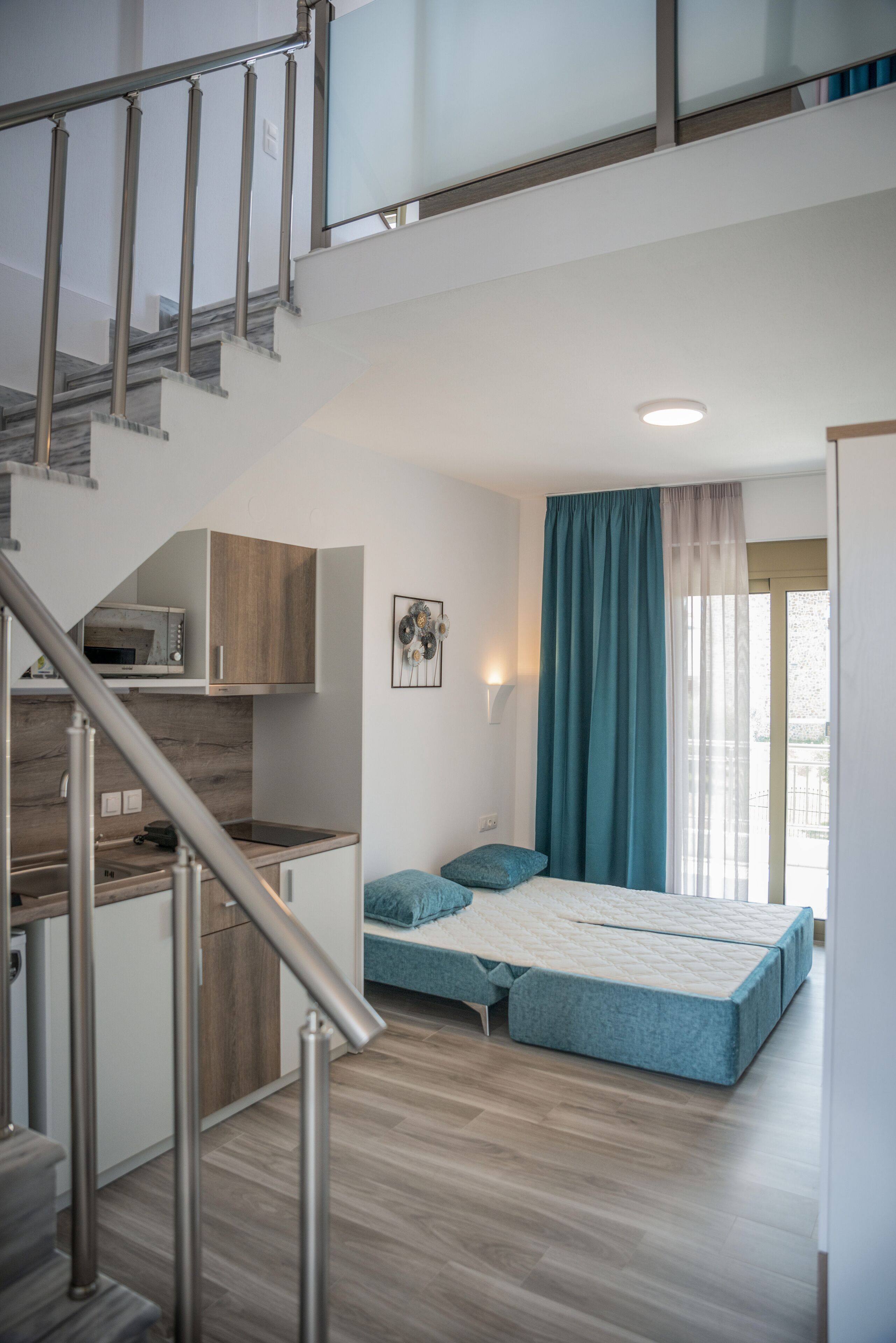 A modern apartment with a kitchen, a bed with a blue mattress, a staircase, and a view of the outdoors.
