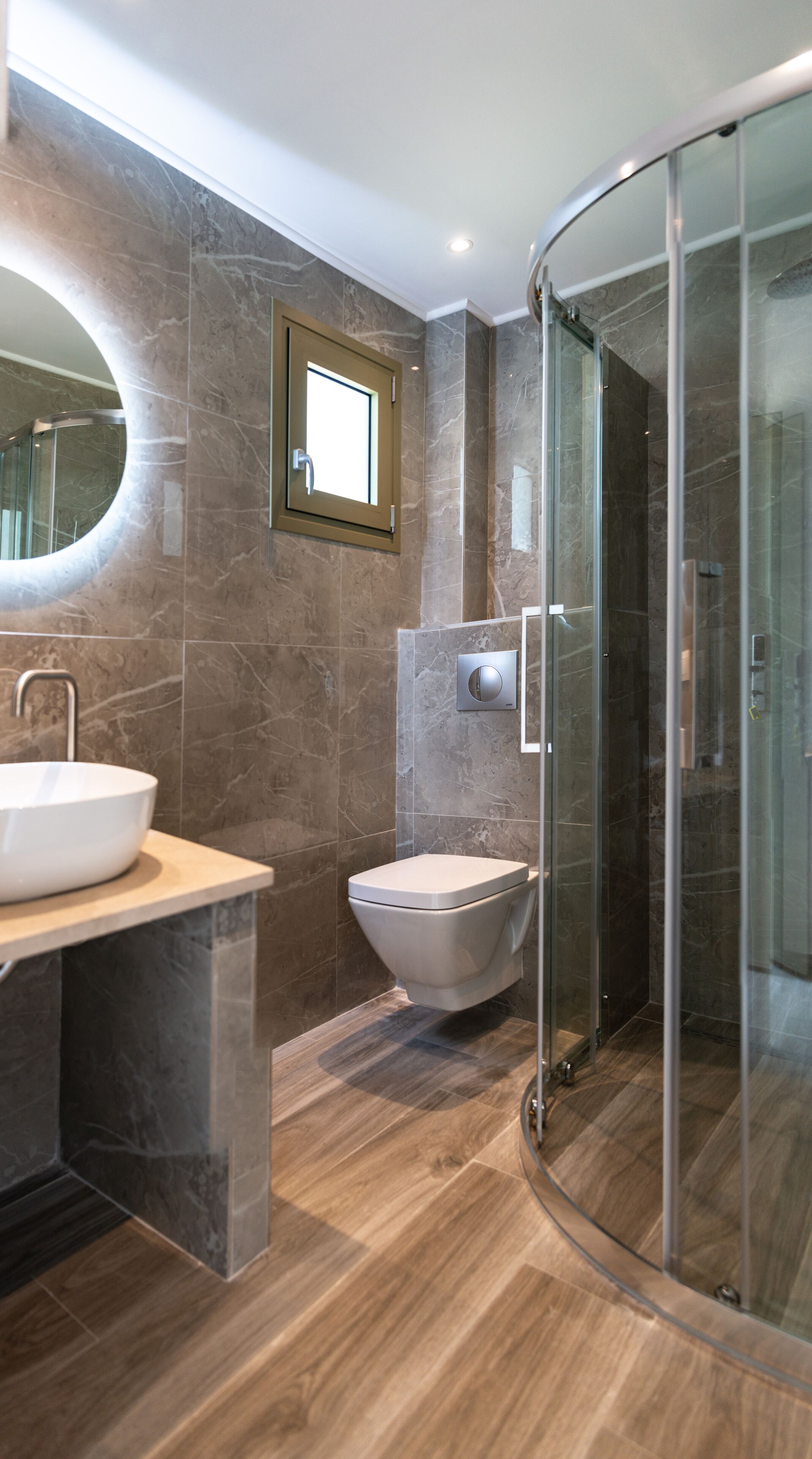 Un baño moderno con un espejo redondo, un lavamanos, un inodoro y una ducha con cerramiento de vidrio.