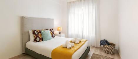 A modern hotel room with a bed, a bedside lamp, a window with curtains, and a small side table.