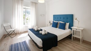 Un dormitorio con cama, una silla, una mesita de noche, una lámpara y una ventana con cortinas.