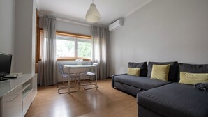 Ein modernes Wohnzimmer mit grauem Sofa, einem Esstisch mit Stühlen, einem Fernseher auf einem Schrank und einem Fenster mit Vorhängen.