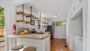 A modern kitchen with white cabinetry, wooden countertops, and a central island with stools. There are hanging shelves above the island and a fire extinguisher mounted on the wall.
