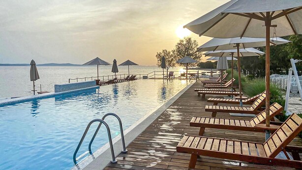 Une piscine à débordement avec des chaises longues en bois et des parasols, offrant une vue sur une mer calme.