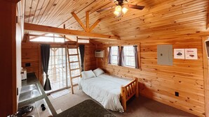 A wooden cabin with a bed, a ceiling fan, a window with curtains, and a kitchenette. - Deluxe Cabin6 Within Campground (Mifflinburg)