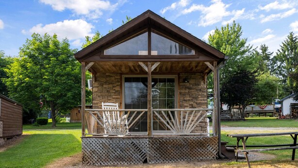 A small wooden cabin with a balcony, stone walls, and a clear view of the surrounding trees. - Deluxe Cabin6 Within Campground (Mifflinburg)