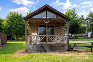 A small wooden cabin with a balcony, stone walls, and a clear view of the surrounding trees. - Deluxe Cabin6 Within Campground (Mifflinburg)