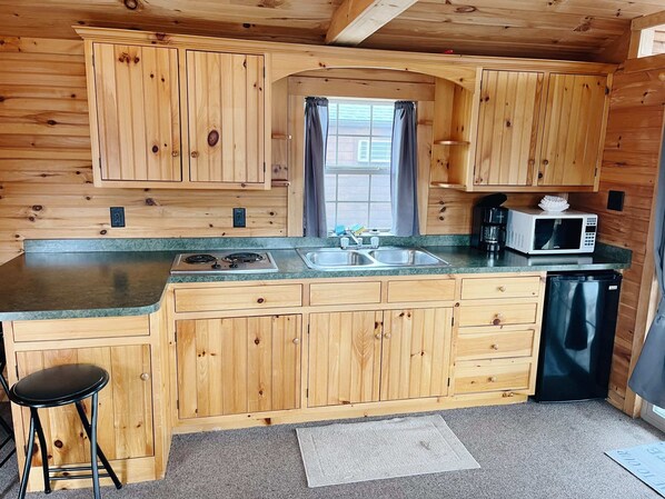 A compact kitchen with wooden cabinets, a double sink, a stove, a microwave, and a refrigerator. - Deluxe Cabin6 Within Campground (Mifflinburg)