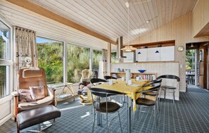Una habitación acogedora y luminosa, con un ventanal grande, una mesa de comedor con sillas, un sofá y una zona de cocina con armarios y una lámpara colgante.