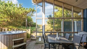 Un patio con jacuzzi de madera, una mesa con sillas y amplias ventanas que ofrecen vistas a árboles y una valla.