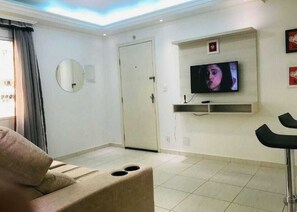 Un salon avec un canapé, un téléviseur fixé au mur, une porte et une fenêtre avec des rideaux.
