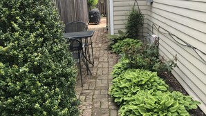 A narrow brick pathway leading to a patio area with a table and chairs, surrounded by lush greenery and a wooden fence. - Oyster Cottage  - walk to shops , Mainstay music venue and the Bay! (Rock Hall)