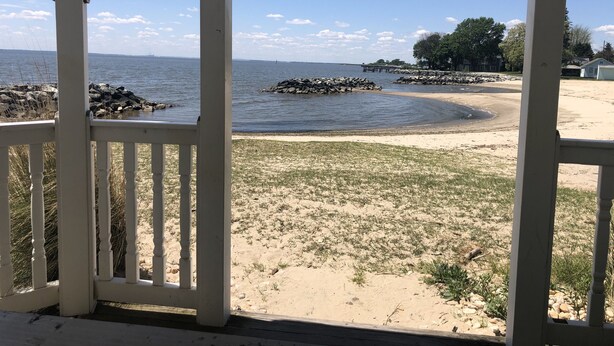 A beach view from a covered area with a wooden deck, sandy beach, rocky shoreline, and a pier in the distance. - Oyster Cottage  - walk to shops , Mainstay music venue and the Bay! (Rock Hall)