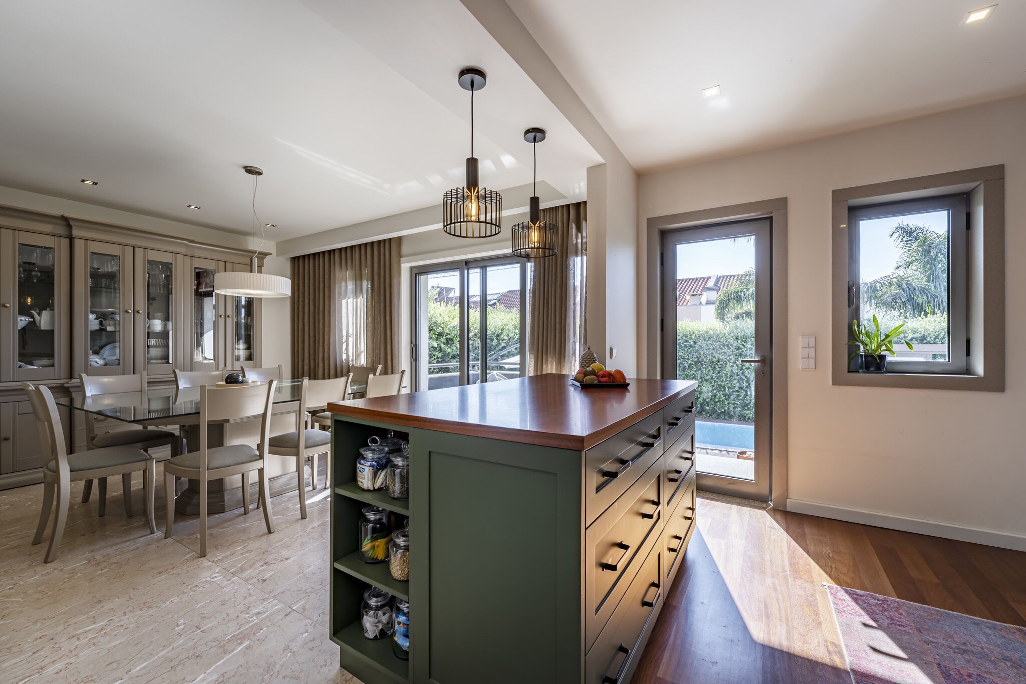A modern kitchen with a central island, dining area with chairs, and large windows offering views of greenery.
