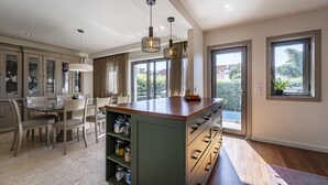 A modern kitchen with a central island, dining area with chairs, and large windows offering views of greenery. - Atlantic House - Beachfront Luxury House (Vila Nova de Gaia)
