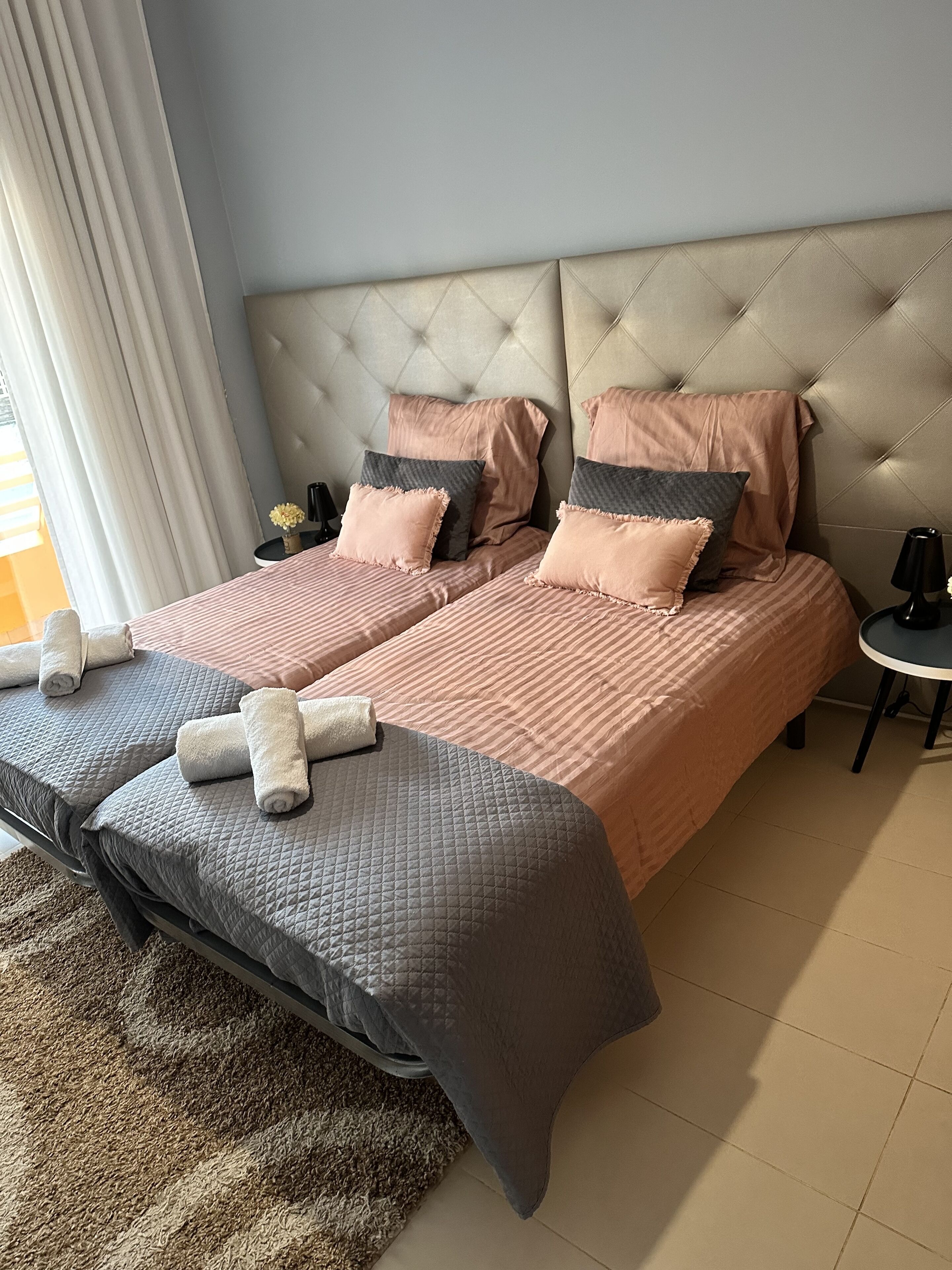 Una cama doble con cabecera capitonada, dos mesitas de noche y un ventanal con cortinas.