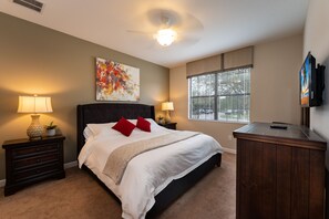 Une chambre à coucher comprenant un lit, deux tables de chevet, une commode, un ventilateur de plafond, une fenêtre avec des stores et un téléviseur fixé au mur.