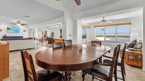 A spacious dining area with a large round table and wooden chairs, a kitchen in the background, and a view of the outdoors through large windows. - Beautiful 5/3 Pool, Saltwater Cove, Sleeps 10! (Saint James City)