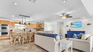 A modern kitchen with wooden cabinets, a dining area with wicker chairs, a sofa, and a television on the wall. - Beautiful 5/3 Pool, Saltwater Cove, Sleeps 10! (Saint James City)