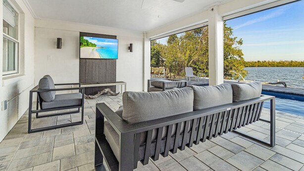 A modern outdoor living area with a sofa, a TV, and a view of a lake. - Beautiful 5/3 Pool, Saltwater Cove, Sleeps 10! (Saint James City)