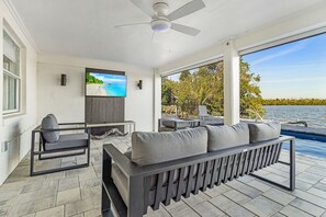 A modern outdoor living area with a sofa, a TV, and a view of a lake. - Beautiful 5/3 Pool, Saltwater Cove, Sleeps 10! (Saint James City)