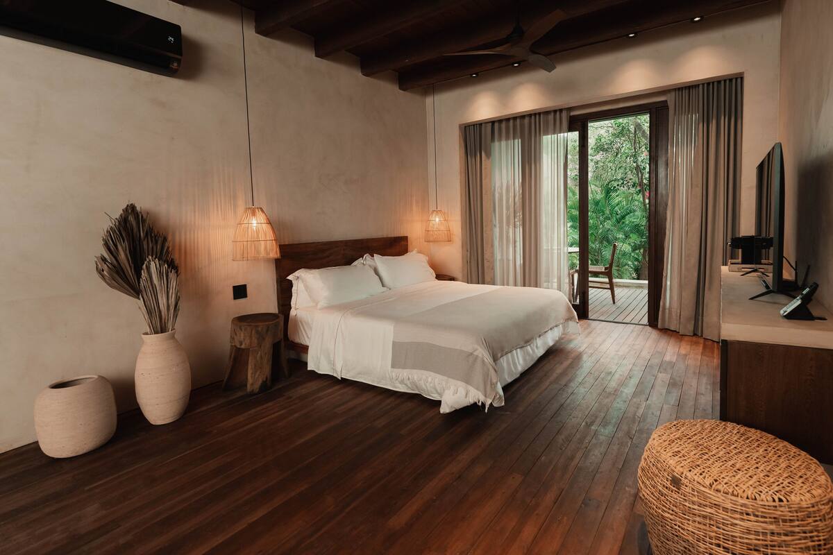 A modern bedroom with a large bed, wooden flooring, a TV, and a view of greenery through the window.