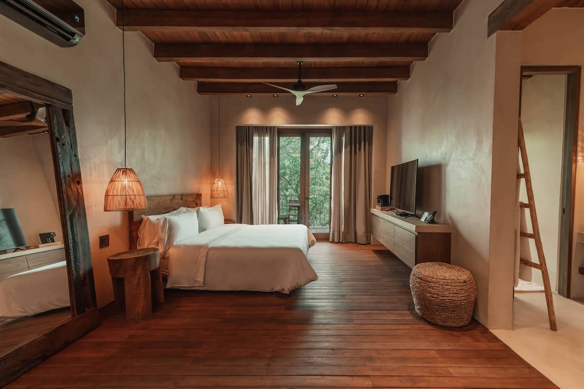 A bedroom with a wooden floor, a bed, a TV, a lamp, and a window with curtains.