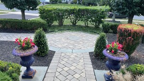 Ein angelegter Garten mit gepflastertem Weg, Blumentöpfen und einem weißen Haus im Hintergrund.