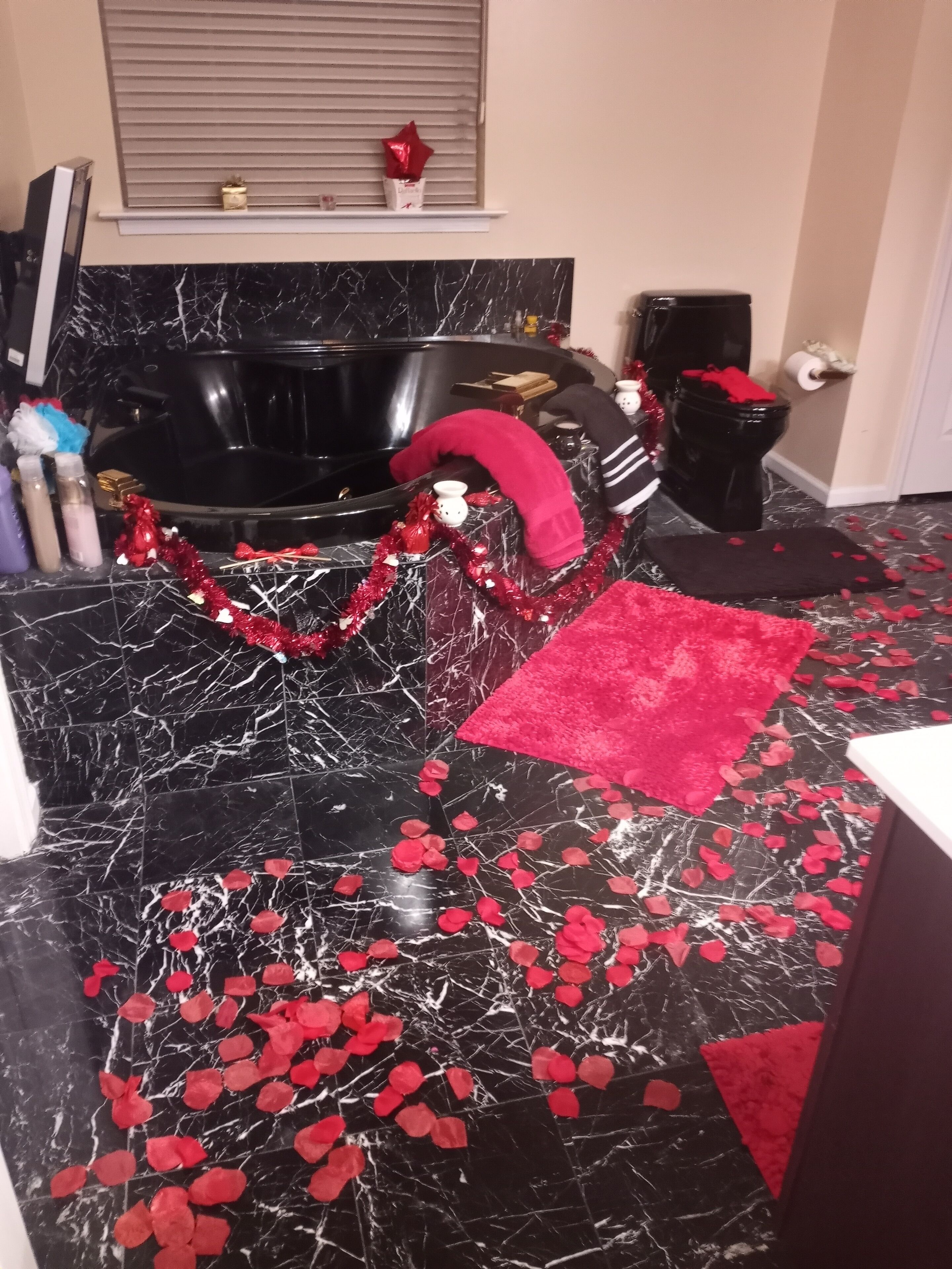 Un baño con piso de mármol negro, tapete de baño rojo y una ventana con persianas.