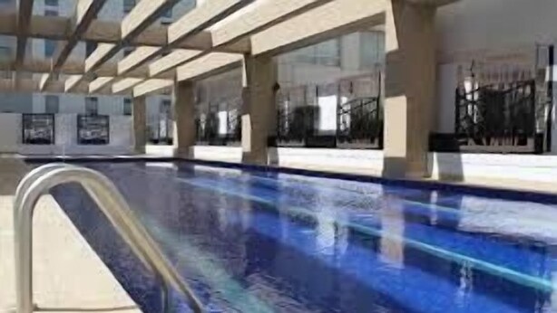 Ein Schwimmbecken mit klarer, blauer Wasseroberfläche, umgeben von einem modernen Bauwerk mit großen Fenstern und einem Metallgeländer.