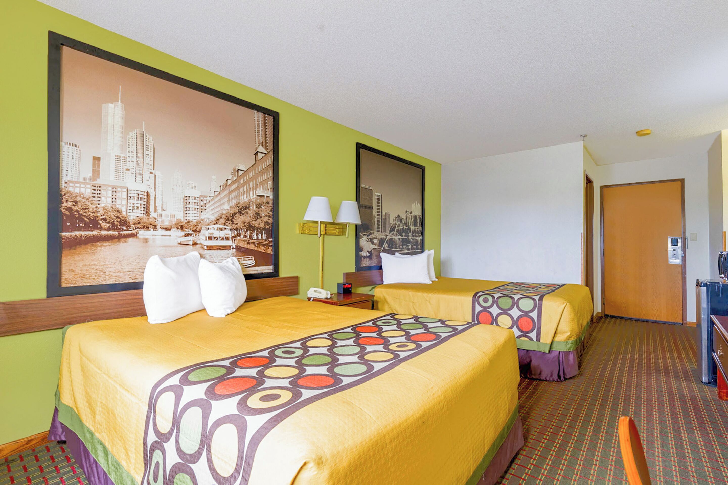 Habitación de hotel con dos camas, un cuadro grande con una vista urbana, un escritorio con lámpara y una puerta de madera.