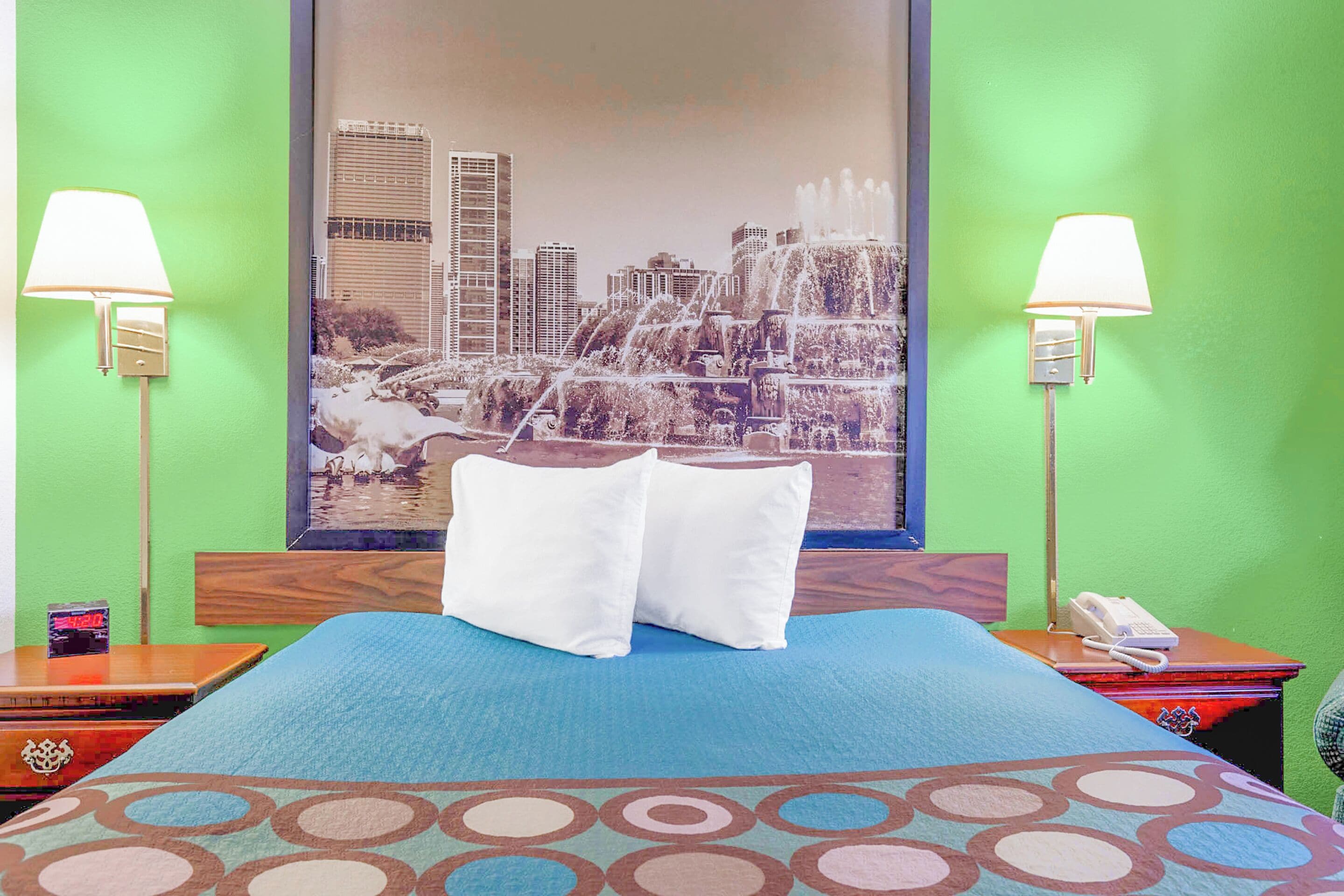 A hotel room with a bed, pillows, a nightstand, a lamp, a phone, and a cityscape painting on the wall.