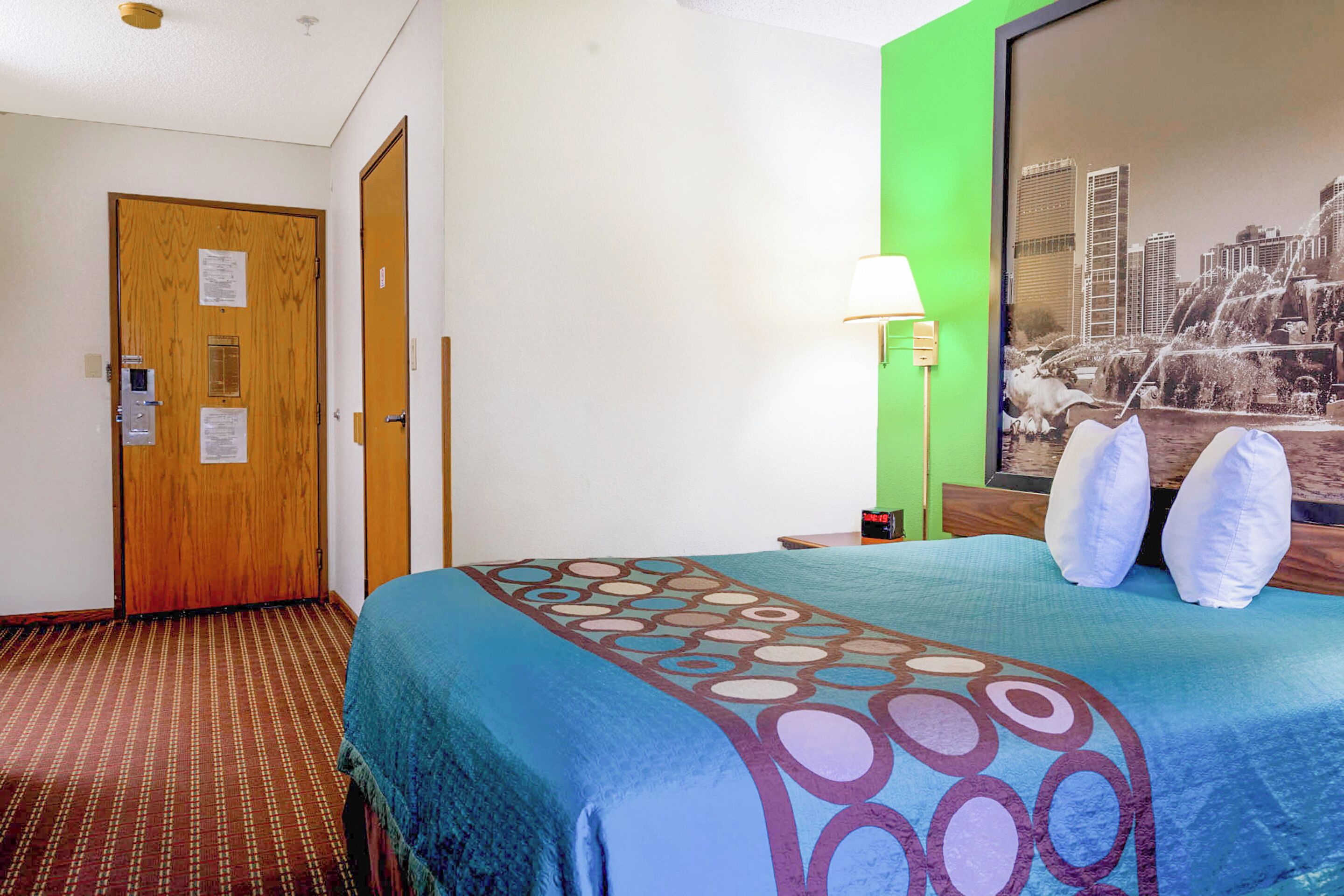 A hotel room with a bed, a green wall, a cityscape painting, and a door with a security panel.