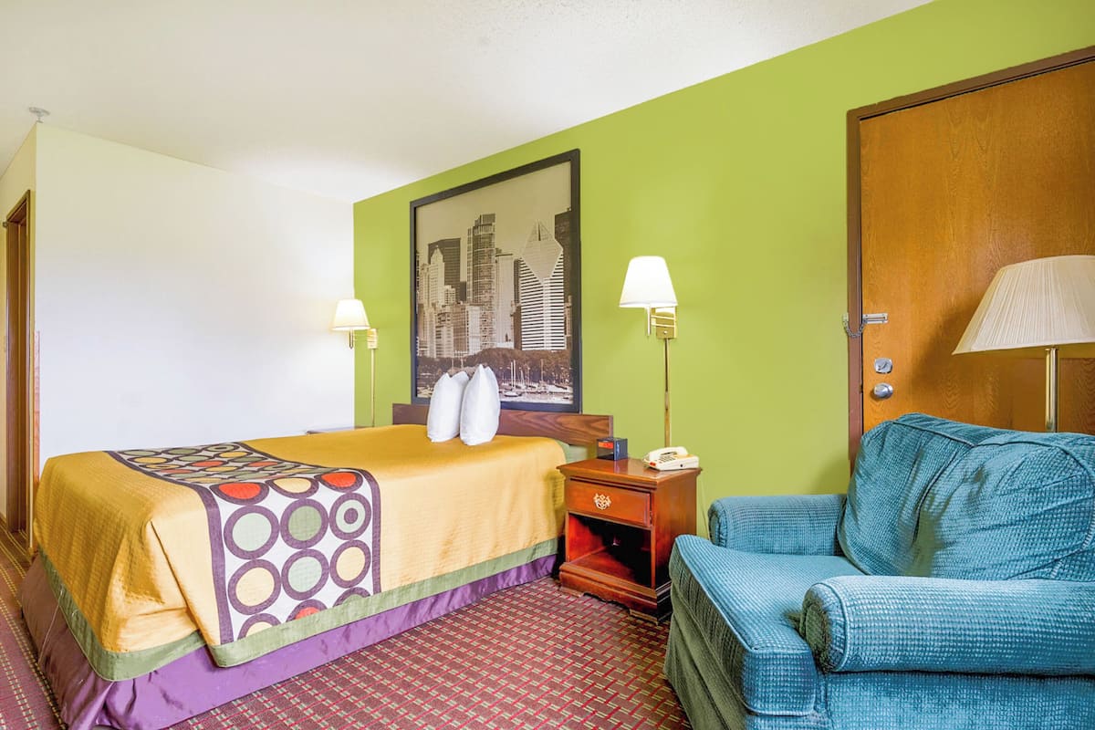 Quarto de hotel com uma cama, uma cadeira, uma mesa de cabeceira, um abajur e um quadro na parede.