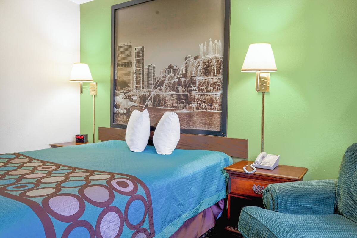 Quarto de hotel com cama, duas luminárias fixadas na parede, telefone e quadro com vista urbana.