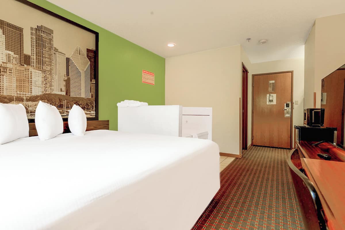 Quarto de hotel com uma cama grande, uma escrivaninha com um computador, uma televisão e uma porta que dá para outro quarto.