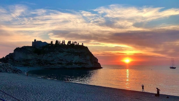 A coastal sunset with a cliff and a building on top, a beach, and a sailboat in the distance. - The Queen Tropea  (Tropea)