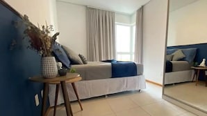 Un dormitorio con cama, una mesita con un florero, un espejo y una ventana con cortinas.
