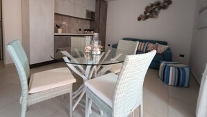 Un soggiorno moderno con un tavolo da pranzo in vetro circondato da sedie in vimini, un divano blu con cuscini decorati e un angolo cottura sullo sfondo.