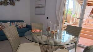 Zona pranzo con tavolo in vetro circondato da sedie in vimini con cuscini. Sullo sfondo, un divano blu con cuscini decorati.
