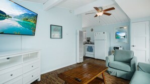A living room with a wooden coffee table, a white entertainment unit, a ceiling fan, and a kitchen area with a refrigerator and microwave.