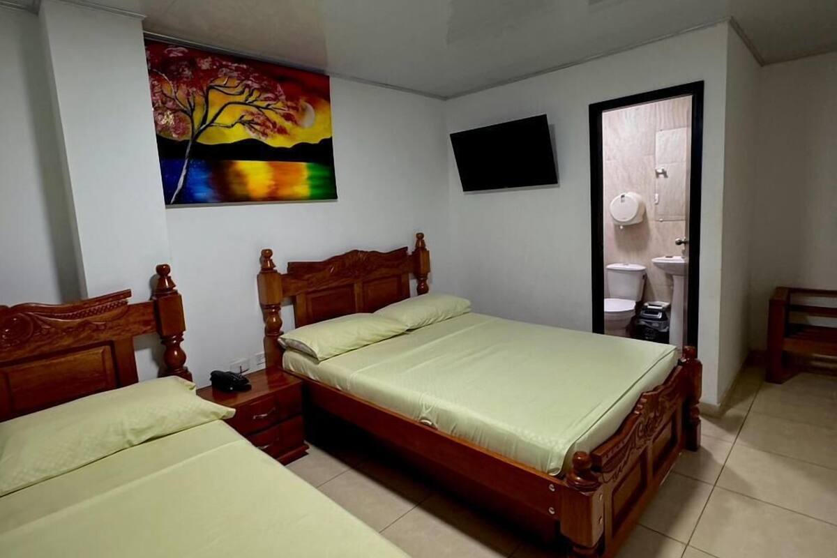 A bedroom with two wooden beds, a ceiling light, a painting, a television, and a bathroom door.