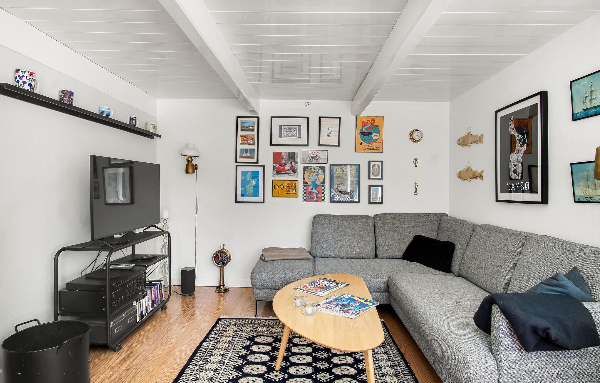 A modern living room with a grey sofa, a wooden coffee table, and a TV on a black stand. There are framed pictures on the wall, a black shelf with decorative items, and a black trash bin.